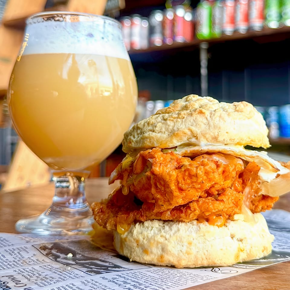 A fried chicken biscuit sandwich with honey and a foamy beer on newspaper at Underground Coffee and Ales.