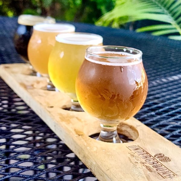 A flight of four craft beers on a wooden paddle, on a black mesh table.