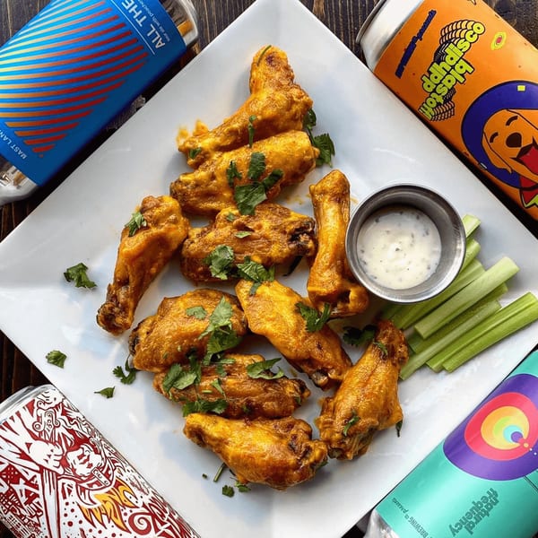 A plate of golden-brown chicken wings with cilantro, ranch, celery sticks, and craft beer cans.