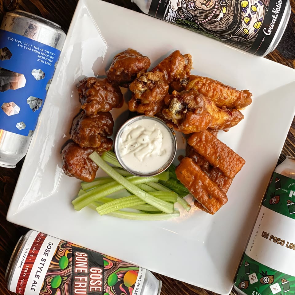 A white plate of saucy wings, boneless bites, celery, ranch, and craft beer cans.
